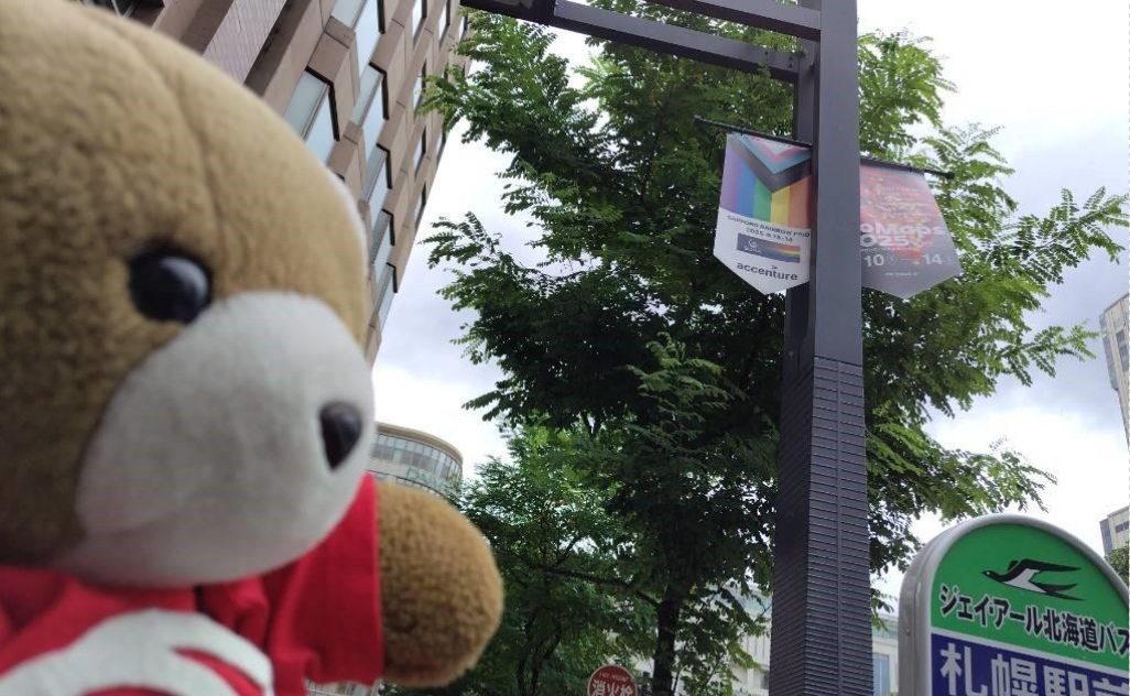 くまちゃんが街路灯を見つめている。 街路灯には、プログレッシブプライドフラッグが掲げられており、旗にはさっぽろレインボープライドのロゴが入っている。