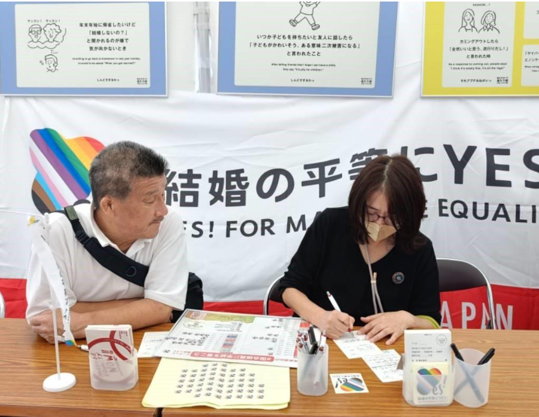 マリフォーブース内の様子。結婚の平等にYES！と書かれた旗が奥にかかっており、その手前のテーブルで2人の参加者が座って、国会議員宛のはがきにメッセージを書いている。