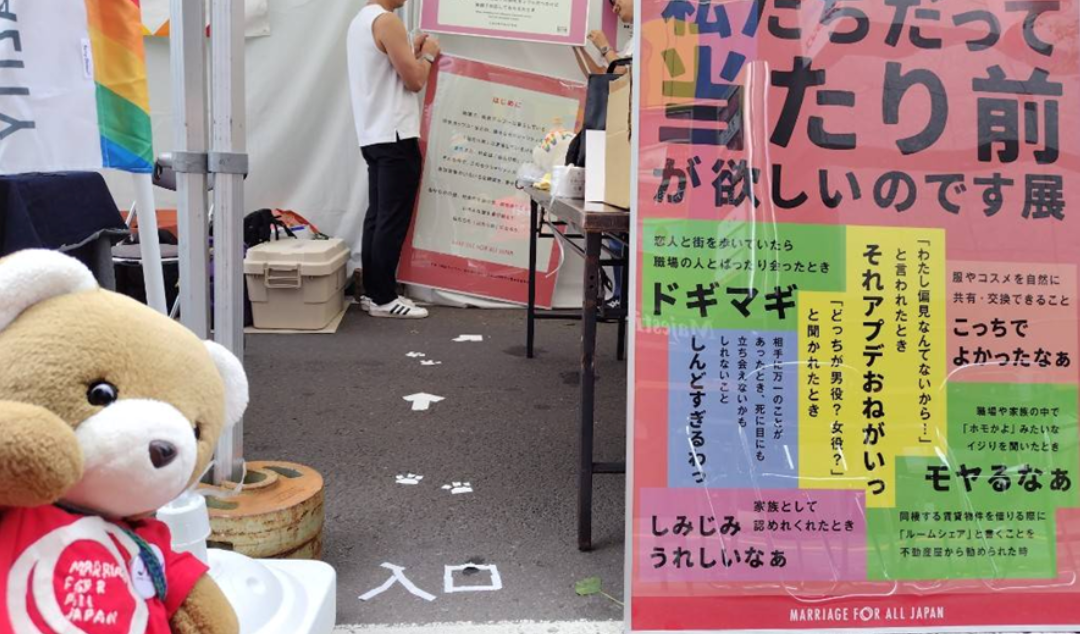 ブース内の展示入口の写真。 「私たちだって当たり前が欲しいのです展」の看板が手前にあり、その横に「入口」という案内が地面に貼られている。地面には、くまちゃんの足跡がついている。 くまちゃんは、どうそいらっしゃいという顔で参加者を招き入れている。