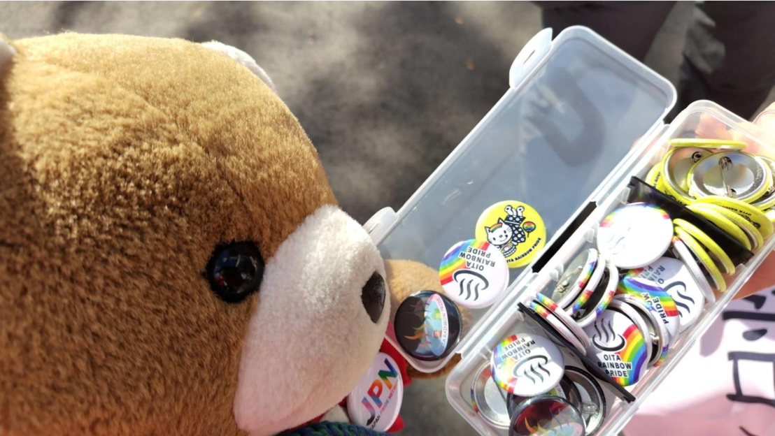 大分レインボープライドのバッヂがたくさん入った箱をながめるくまちゃん
