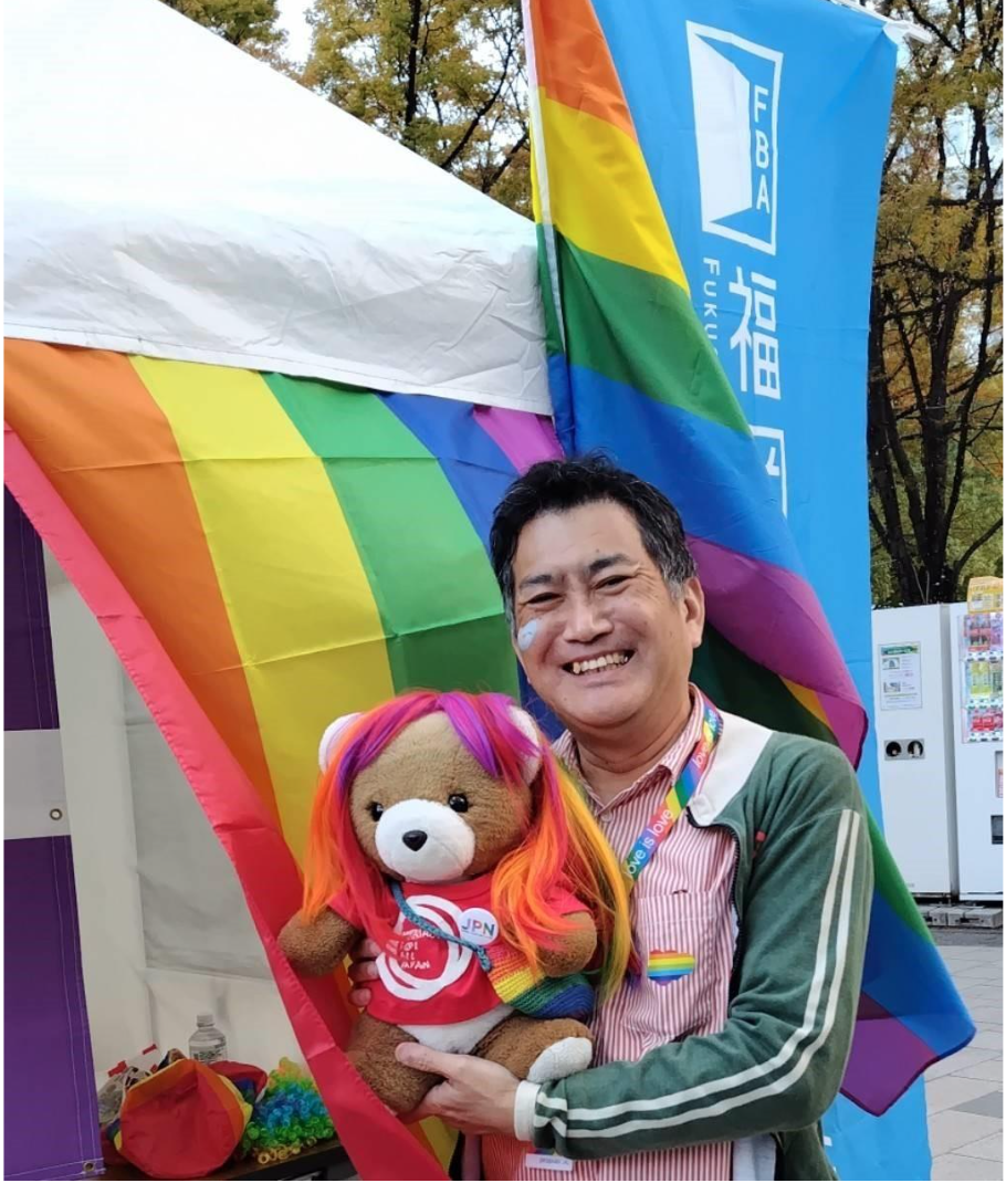 福岡県弁護士会の旗とレインボーフラッグが飾られたブースの前に弁護士が立っている。 弁護士がくまちゃんを抱いていて、くまちゃんはウイッグをかぶっている。