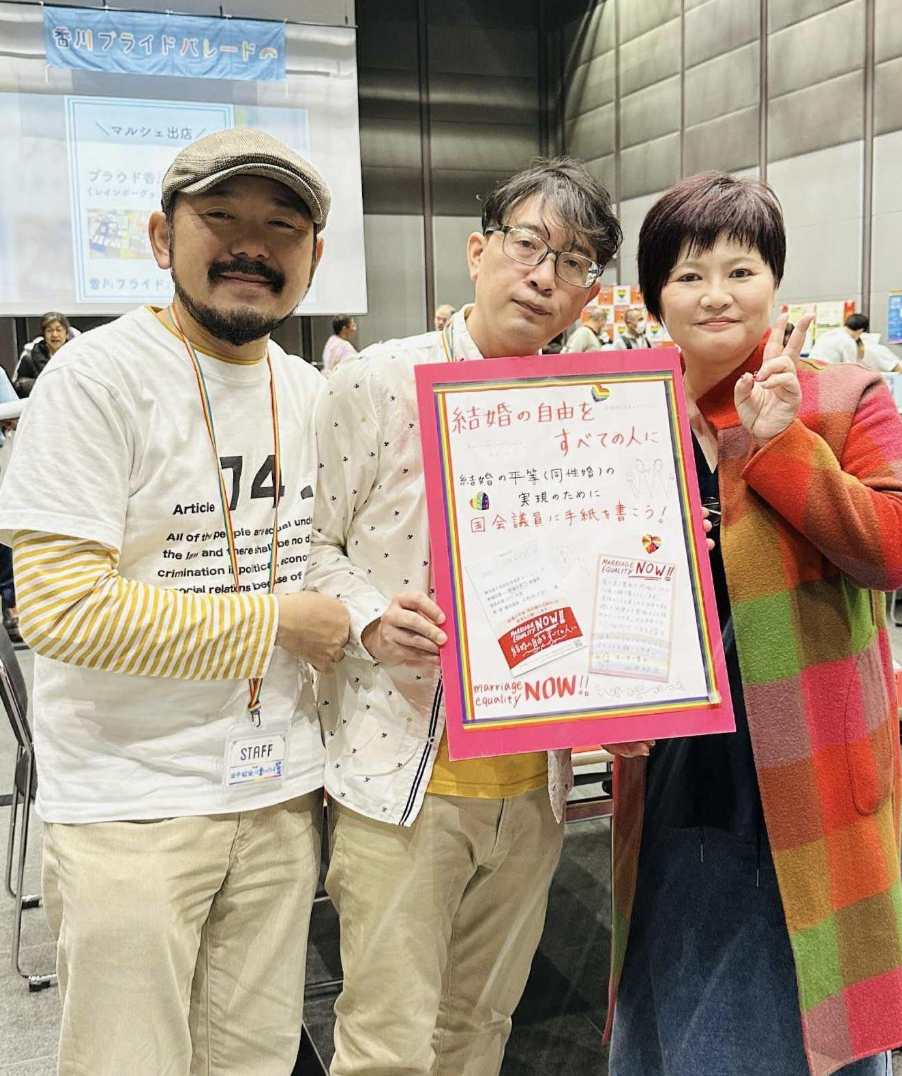 香川プライドパレードのマルシェ会場の様子。 会場で、３人の人物が記念撮影しており、左二人は原告カップルで、国会議員へ手紙を書こうと呼びかけるボードを掲げている。右側の人物は弁護団の弁護士で、カラフルなコートに身を包んでいる。