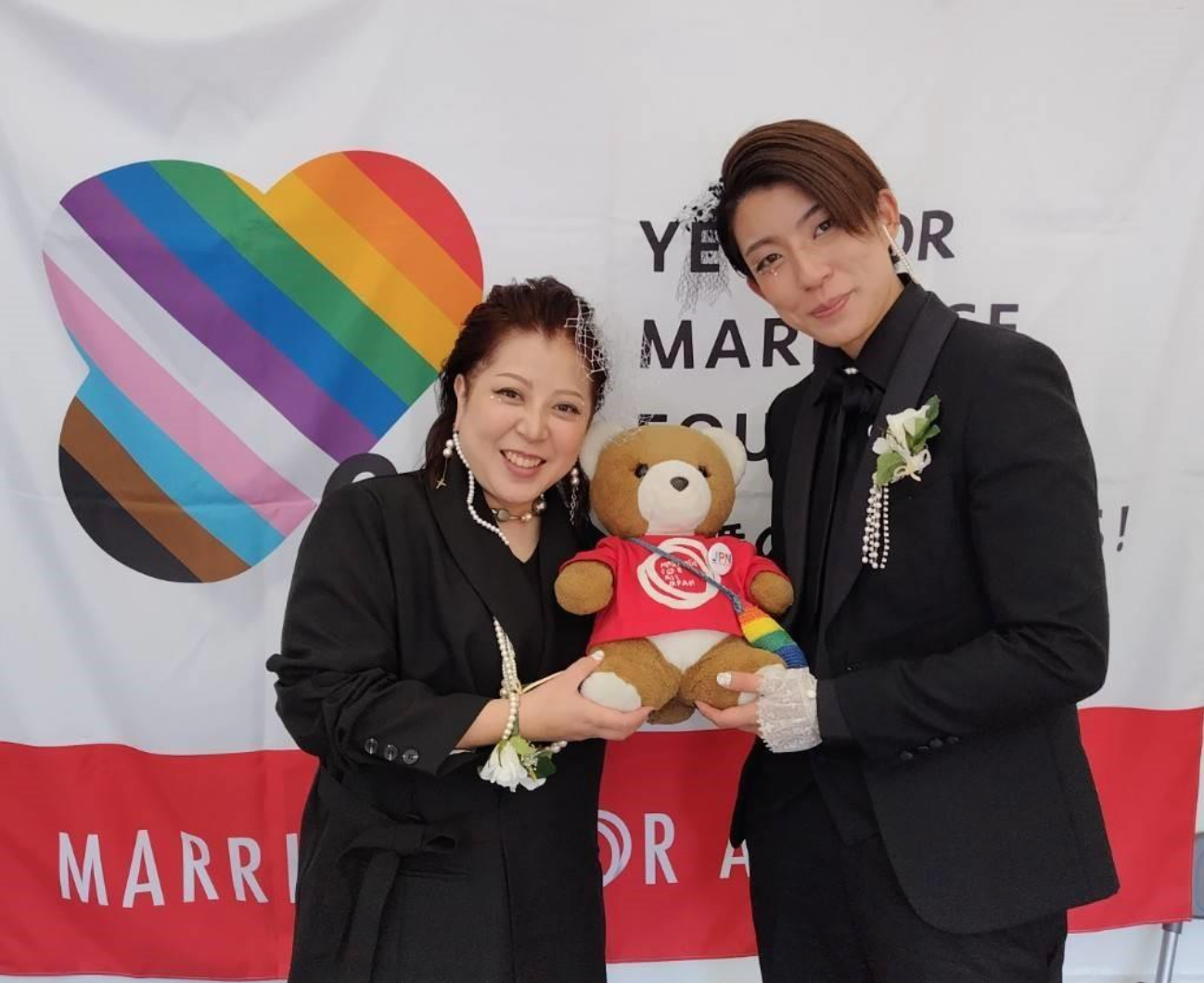 ウエディング衣装をきたカップルが二人で並んでおり、その間にマリフォ―くまちゃんがいる。