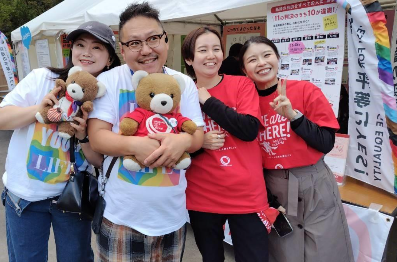 マリフォーブース前に４人の人が並んでいる。 中央の人物がくまちゃんを抱いている。 右側の二人はマリフォーのシャツをお揃いで着ている。