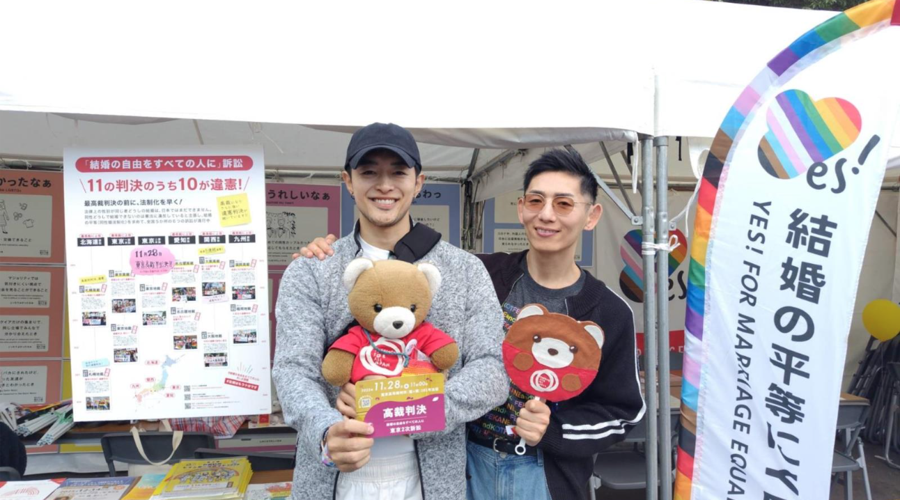 マリフォ―ブース前に二人の男性が立っている。人にがもう一方の肩に他をまわし、仲良さそうにほほえんでいる。 ふたりはそれぞれ、マリフォ―くまちゃんや結婚の自由をすべての人に訴訟のアピールグッズなどをもっている。