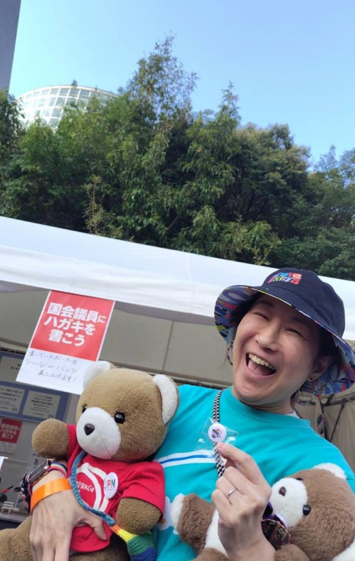 マリフォ―ブース前でくまちゃんを抱く小野春さん。 小野春さんはにじいろかぞくのシャツを着ていて、楽しそうに笑っている。