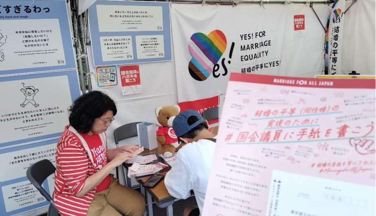 マリフォーブース内のテーブルに、マリオースタッフと参加者が座っている。 参加者は手紙を書いていて、スタッフは手紙の書き方の説明をしている。 そのそばにくまちゃんがいて、お手伝いをしている。