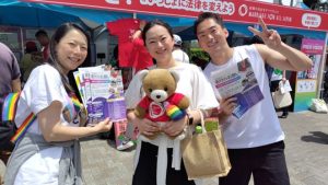 マリフォーブース前で、三人の弁護団メンバーが笑顔で並んでいる。くまは真ん中のひとにだっこしてもらっている。