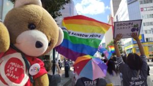さっぽろレインボープライドのパレードを歩くくまちゃん。くまちゃんのすぐそばで、さっぽろレインボープライドの旗がゆらめいている。