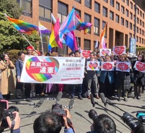 名古屋の裁判所の前で弁護団や支援者が旗を出している。その姿を多くの報道陣が撮影している。 旗には高裁でも違憲と書かれていて、くまも旗を持っている。