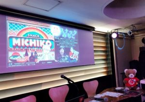 イベント会場のスクリーンに、スナックみちこと書かれたイラストが映し出されている。 そのそばに登壇者用のテーブルがあり、くまがテーブルの上でイベントの準備をしている。