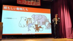 三浦弁護士が壇上でスライドを見せて話をしている。 頼もしい動物のイラストと、いざ最高裁へという言葉が並んでいる。