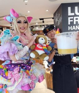 LUSHの店舗内で、ドラァグクイーンのライラと、くまちゃん、店員さんが笑顔で並んでいる。